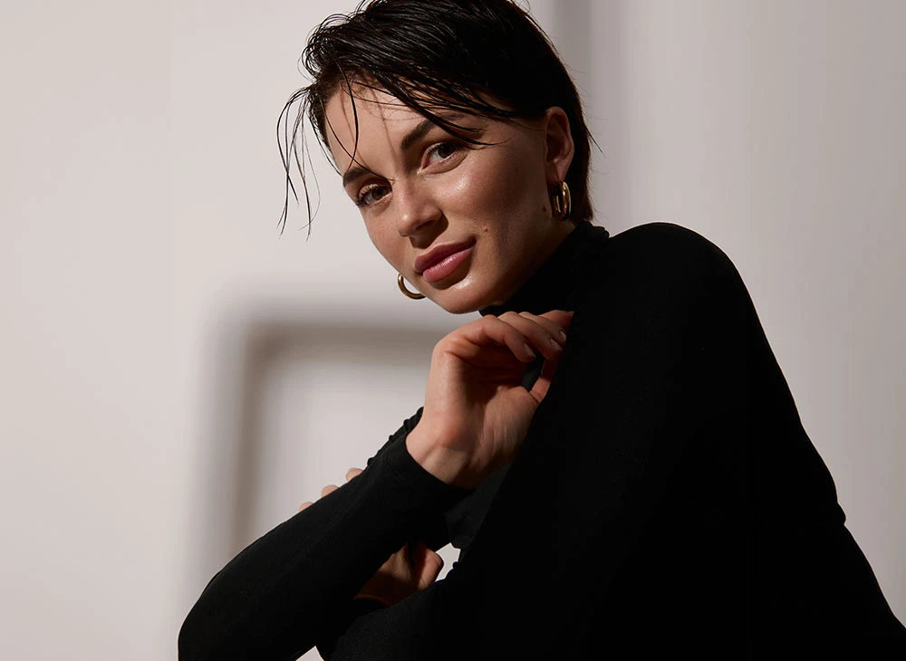 A brunette woman in a black turtle neck with gold jewelry, smiling - Revision Rhinoplasty Procedure in Toronto, Canada