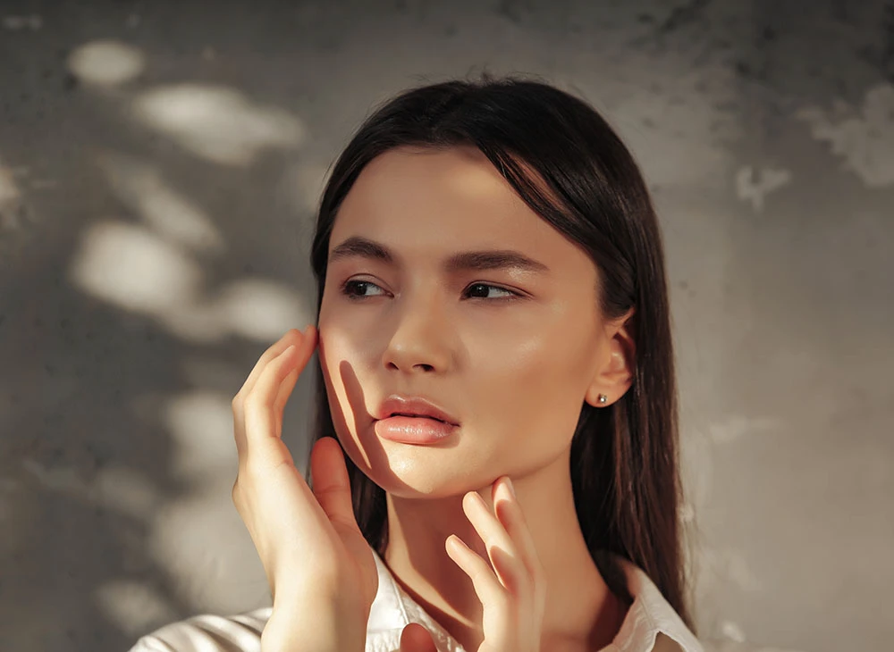 A brunette woman looking to the side, light cast on her face, hands held up to her face - Rhinoplasty Procedure in Toronto, Canada