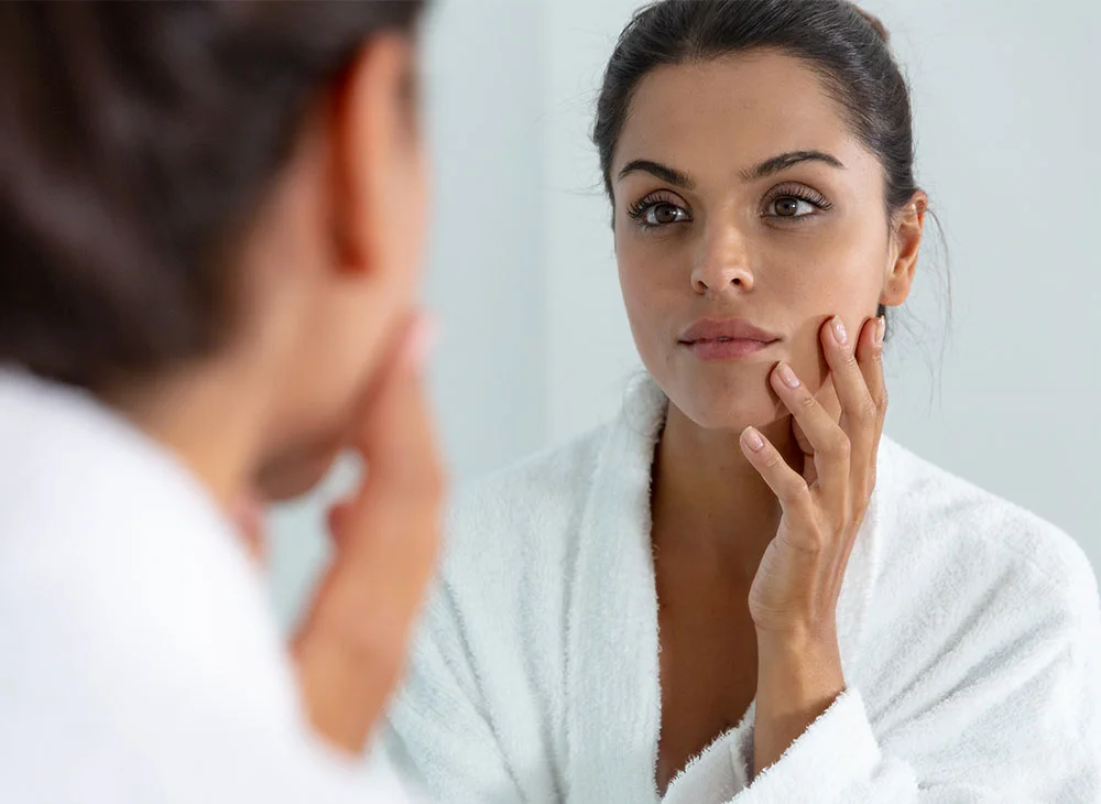 Woman in a white robe touching her face gently while looking at her reflection in the bathroom mirror - Rosacea Treatment in Toronto, ON