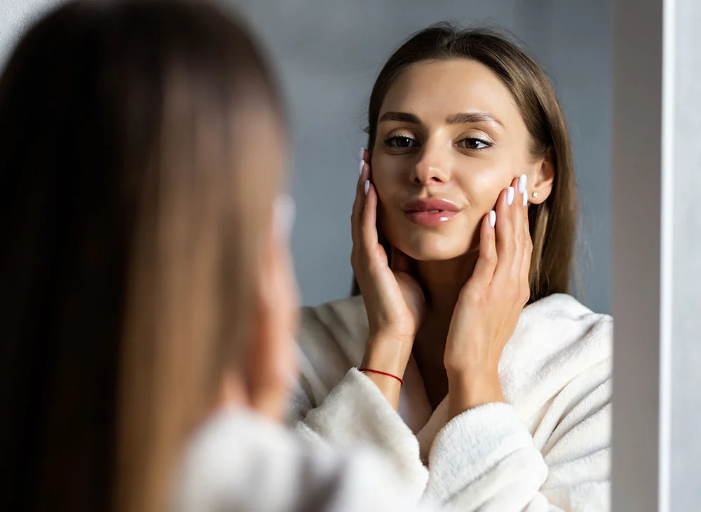 A young woman in a white robe gently touching her face while looking at her reflection in the mirror - Skin Rejuvenation Treatment in Toronto, ON